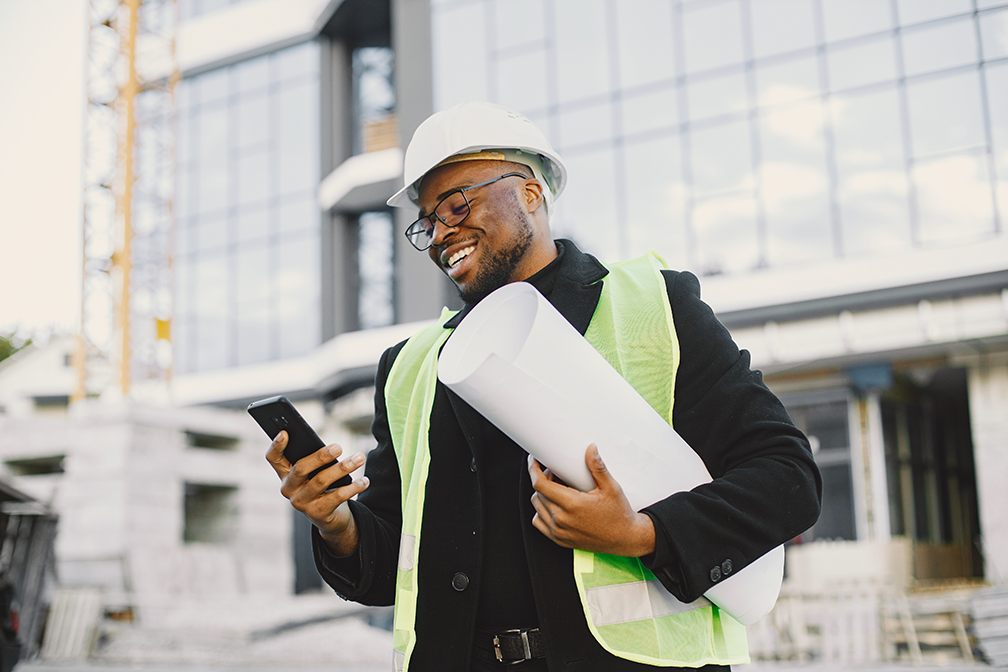 Ingénieur souriant en casque et gilet de sécurité, plans sous le bras, devant un bâtiment en construction